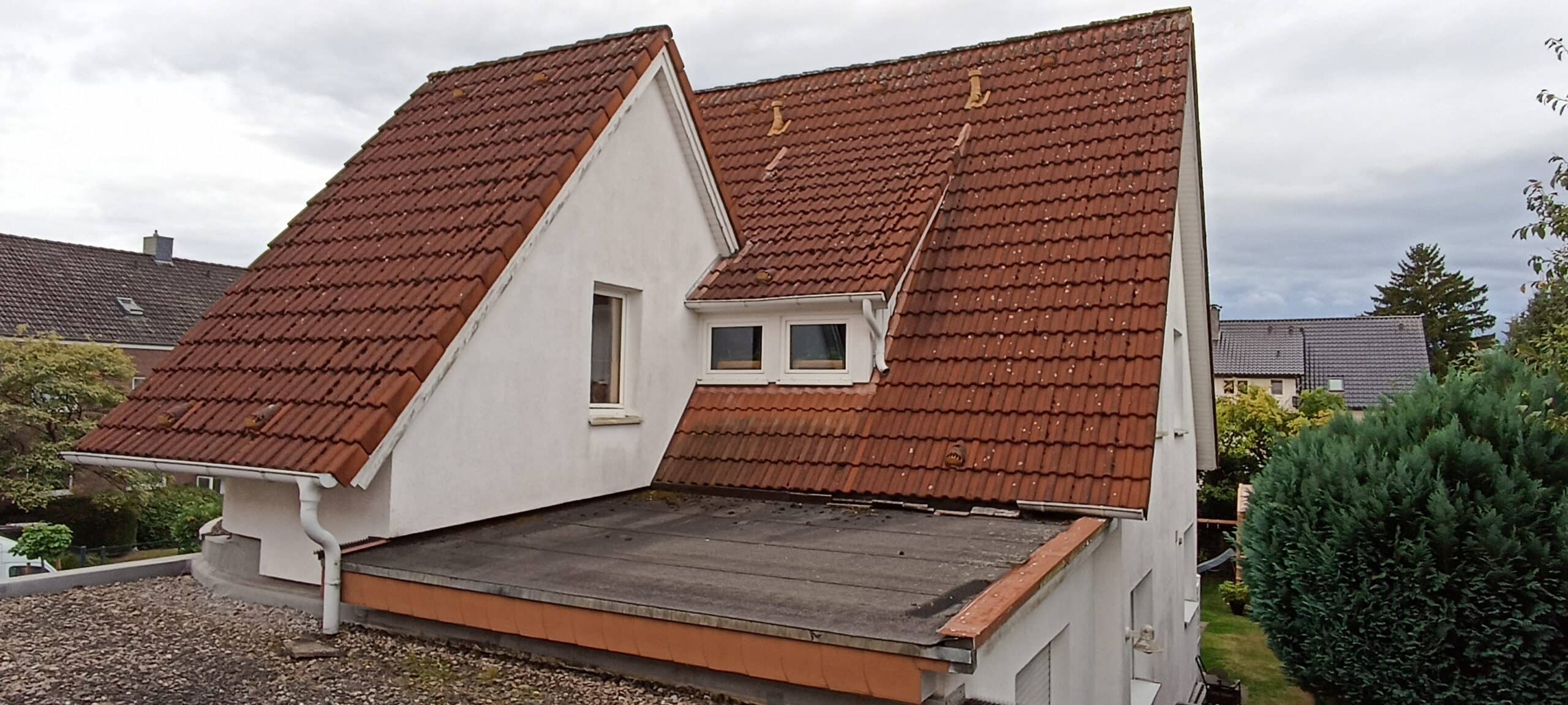 Dach eines weißen Hauses mit roten Ziegeln und einem Flachdachteil, umgeben von Bäumen und weiteren Gebäuden im Hintergrund.