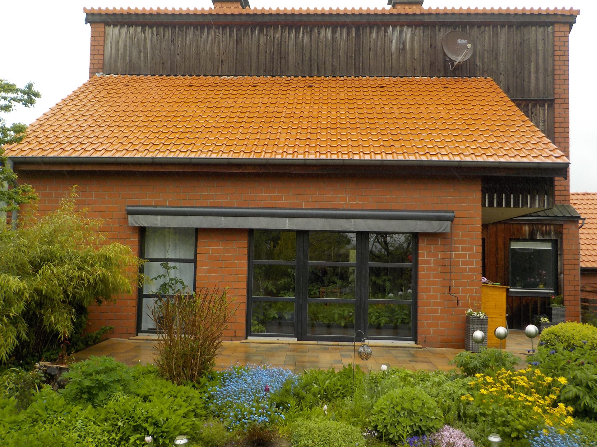 Rotes Backsteinhaus mit orangefarbenem Ziegeldach, großer Terrasse und üppig bepflanztem Garten im Vordergrund.