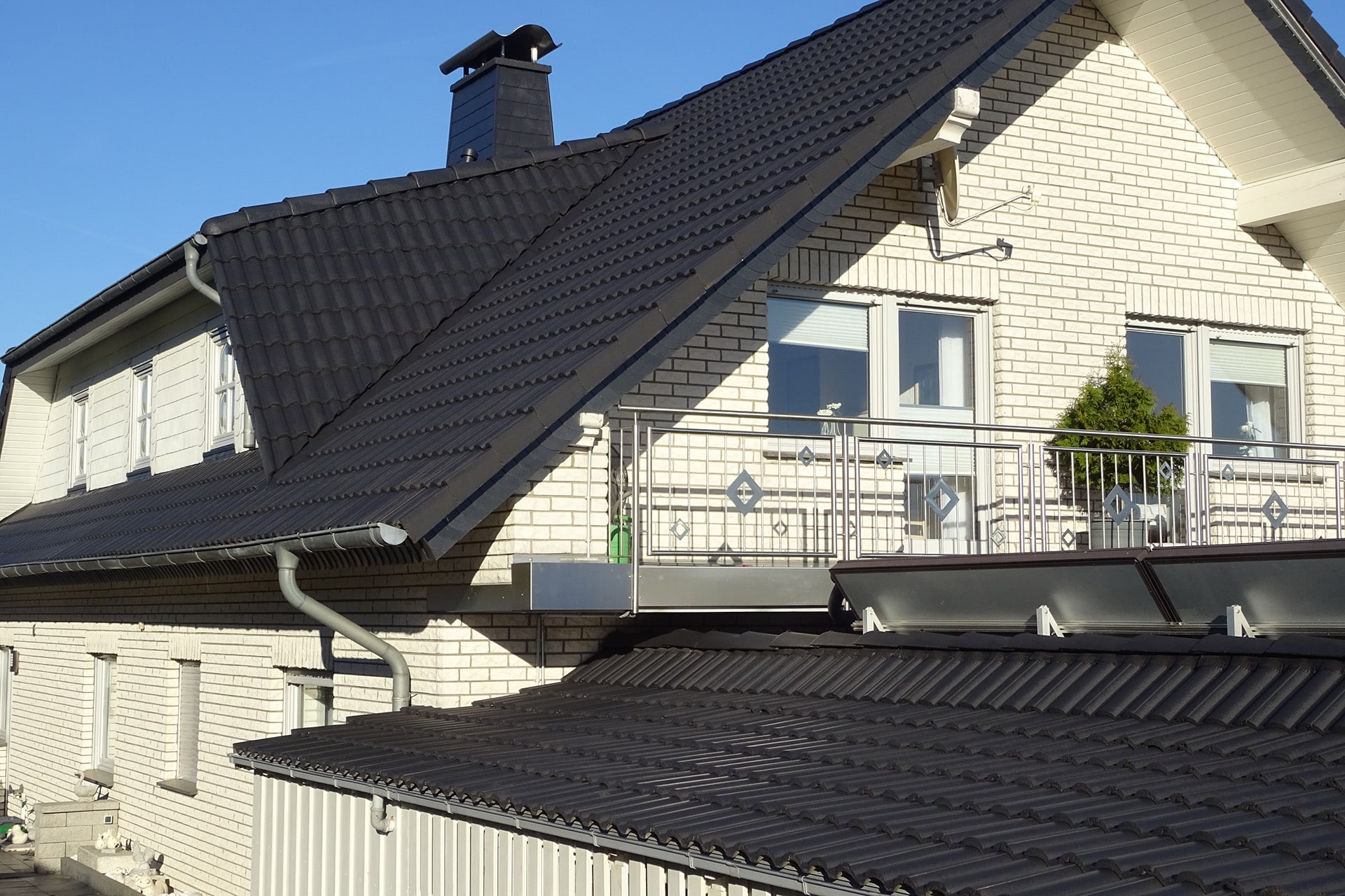 Dach eines hellen Backsteinhauses mit schwarzen Ziegeln, einem Schornstein und einem Balkon mit Glasgeländer.