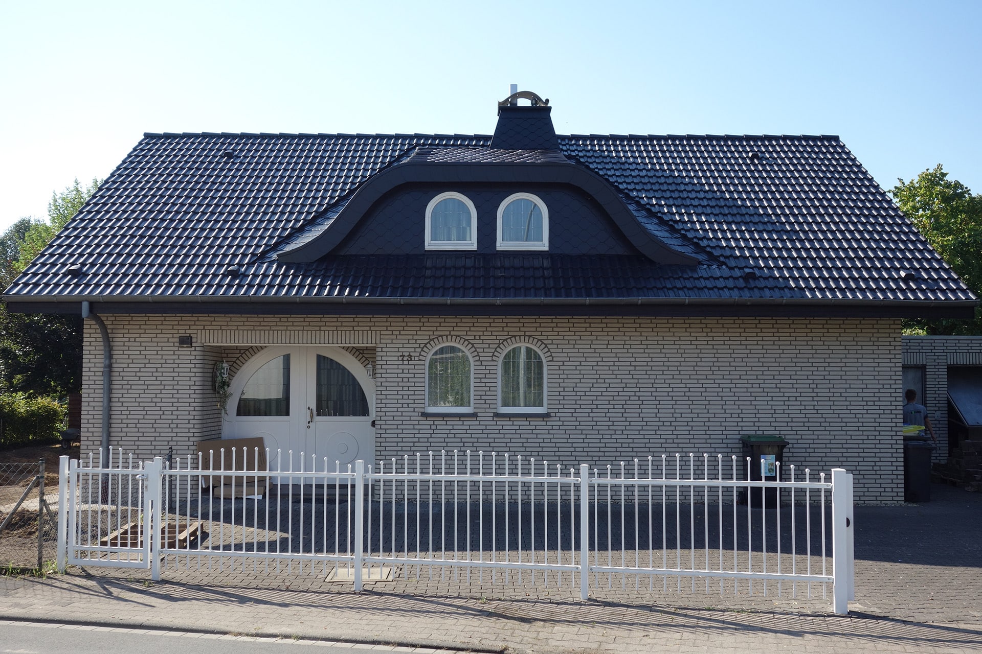 Ein graues Backsteinhaus mit geschwungenem Dach, kleinen Gaubenfenstern und einem weißen Zaun davor.