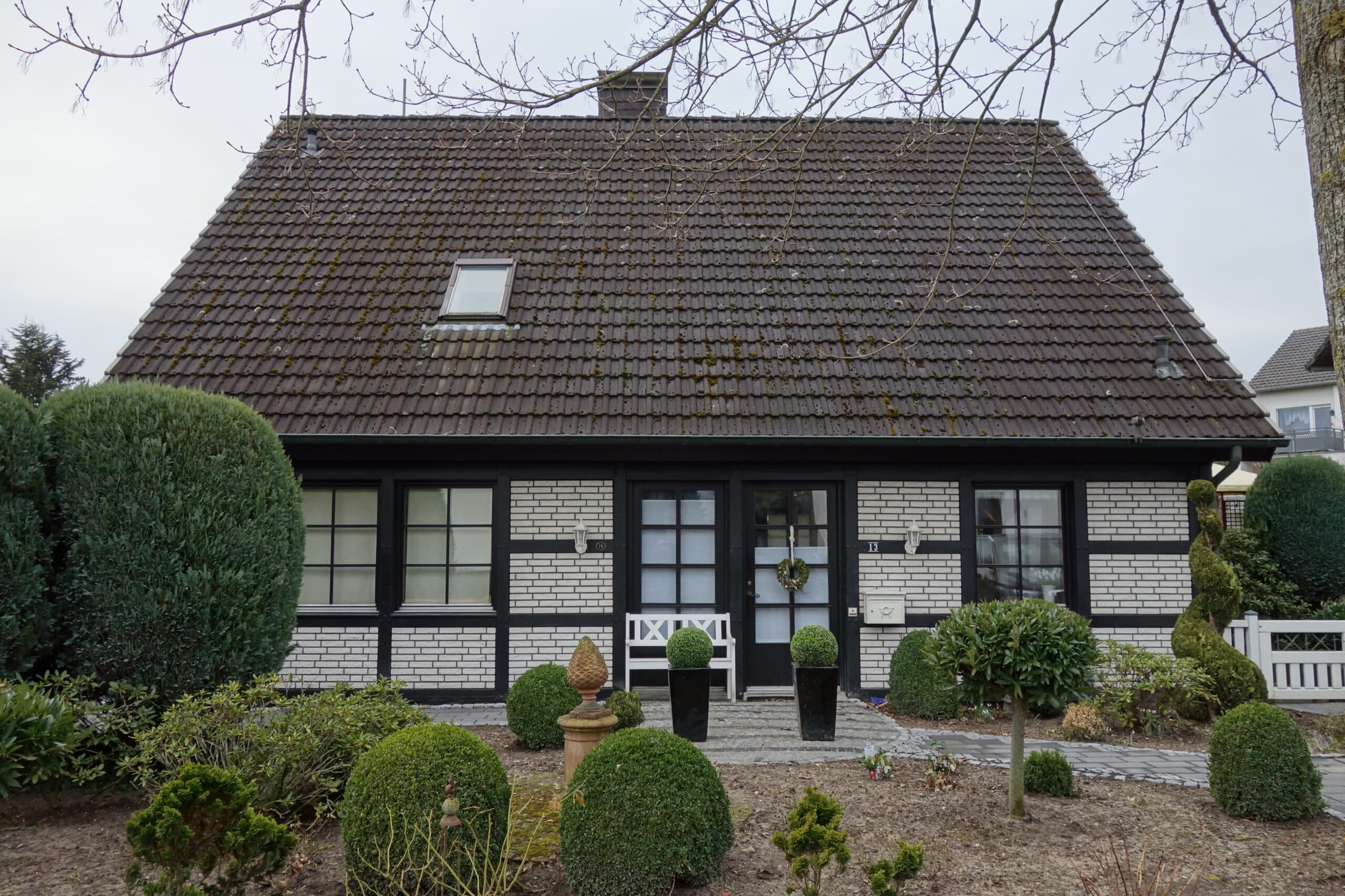Fachwerkhaus mit dunklem Dach und weißer Fassade, umgeben von gepflegtem Garten mit Formschnittbüschen und Hecken.