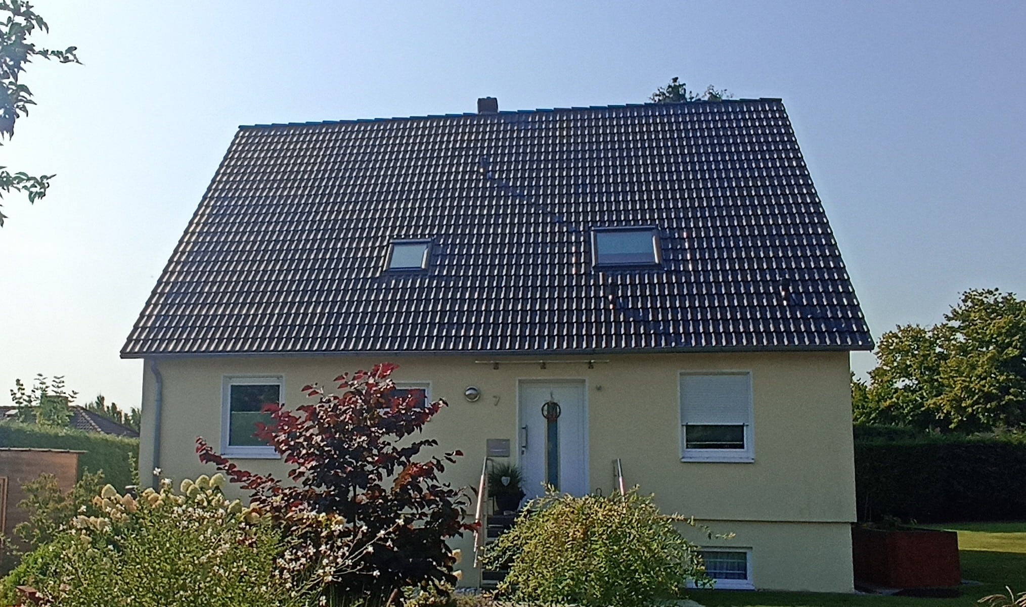Gelbes Einfamilienhaus mit schwarzem Walmdach, zwei Dachfenstern und umgeben von einem Garten mit Sträuchern.