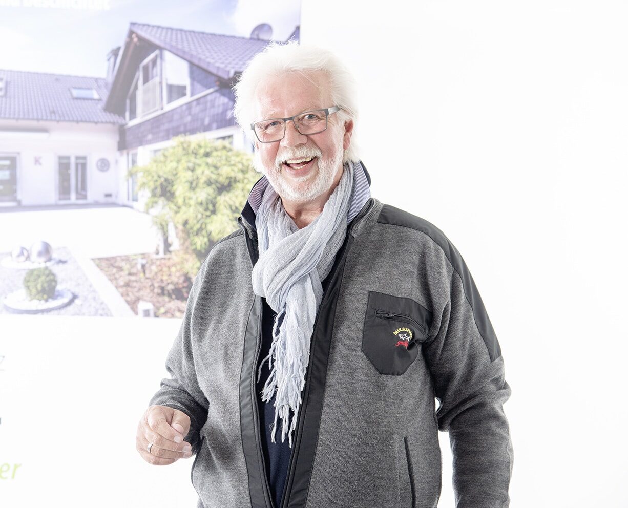 Älterer Mann mit Brille und Schal lächelt vor einem Poster eines Hauses, Logo von "Bautenschutz Bogumil GmbH" auf der Jacke.